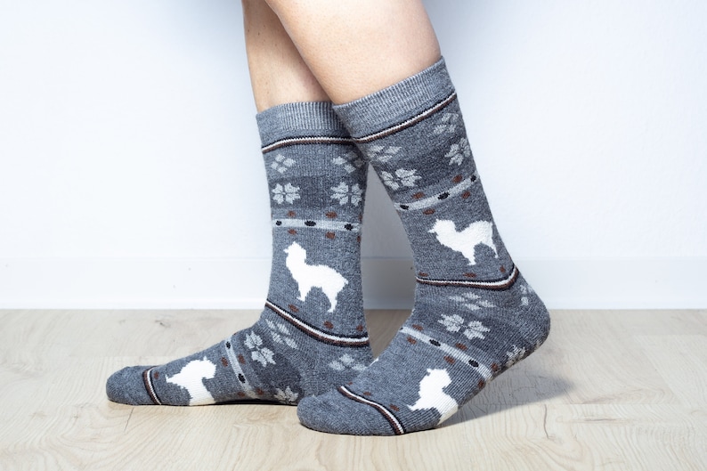Pu&ograve; includere: Un paio di calzini grigi con un motivo di alpaca bianco. I calzini hanno una striscia marrone intorno alla parte superiore e un motivo a fiocco di neve bianco.
