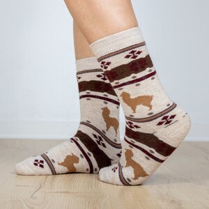 Pu&ograve; includere: Un paio di calzini beige con un motivo marrone e bordeaux che presenta una silhouette di alpaca. I calzini sono realizzati in un materiale morbido e confortevole.