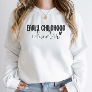Może przedstawiać: Biała bluza z napisem "Early Childhood Educator" czarnymi literami i małym czarnym sercem.