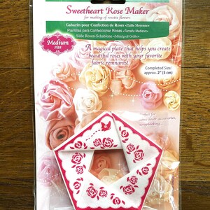 Könnte beinhalten: Eine weiße Plastik-Schablone mit einem rosafarbenen Rosenmuster. Die Schablone wird verwendet, um Stoffrosen zu erstellen. Die Verpackung lautet "Sweetheart Rose Maker" und "Fertige Größe: ca. 2" (5 cm)".
