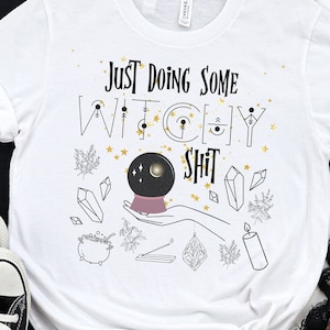 May include: White t-shirt with the text "Just Doing Some Witchy Shit" in black and gold lettering. The design includes a crystal ball, stars, and other witchy symbols.