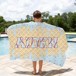 May include: A blue and white waffle-patterned beach towel with a blue and red dripping ice cream design. The towel has the name "Aiden" written in white letters with red baseball stitching.