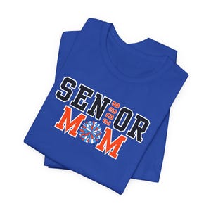 K&ouml;nnte beinhalten: Ein k&ouml;nigsblaues T-Shirt mit dem Aufdruck "SENIOR 2026 MOM" in Schwarz und Orange. Das Design beinhaltet ein Cheerleader-Pompon. Das T-Shirt ist gefaltet.