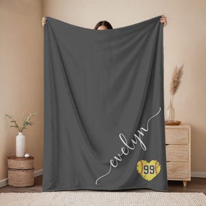 May include: A dark gray blanket with the name "evelyn" in white script. A yellow heart with the number "99" is on the lower right. The blanket is held up by a person.