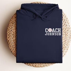 May include: A navy blue hoodie with a drawstring hood, featuring the text "COACH JOHNSON" and a baseball graphic embroidered on the front. The hoodie is folded and placed on a woven basket.
