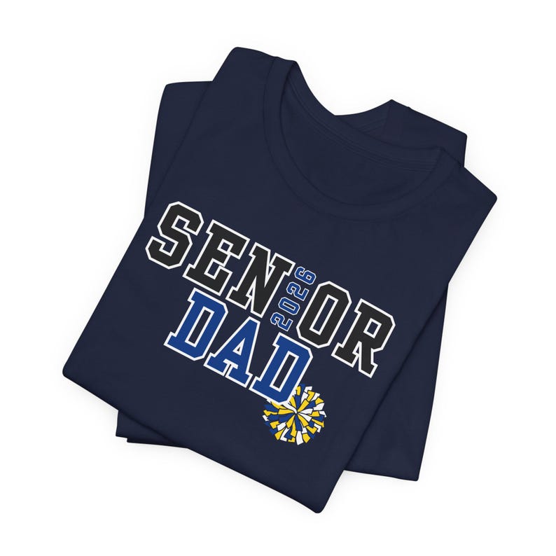 K&ouml;nnte beinhalten: Marineblaues T-Shirt mit dem Text "SENIOR DAD 2023" in wei&szlig;er, schwarzer und blauer Schrift. Eine gelb-blaue Pompon-Grafik befindet sich darunter. Das T-Shirt ist gefaltet.