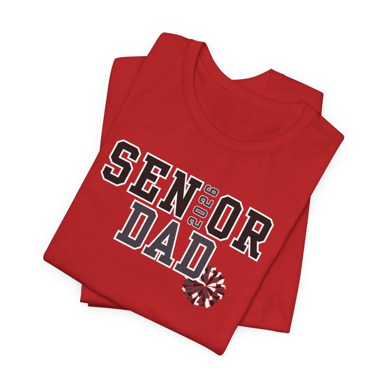 K&ouml;nnte beinhalten: Ein rotes T-Shirt mit dem Text "SENIOR DAD 2023" in schwarzen und wei&szlig;en Blockbuchstaben. Unter dem Text befindet sich eine kleine Grafik eines Cheerleader-Pompons. Das T-Shirt ist gefaltet.