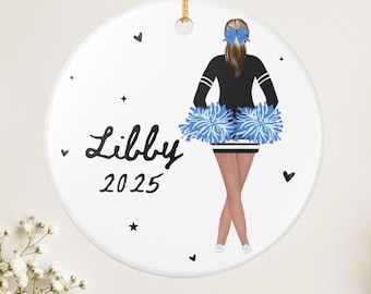 May include: A white ceramic ornament with a gold string for hanging. The ornament features a cheerleader illustration with blue pom-poms, a black and white uniform, and the name "Libby" with the year "2025". Small black hearts and stars are scattered around the design.