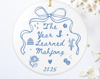 Könnte beinhalten: Weißer Keramikschmuck mit blauem Design und goldener Schnur. Der Schmuck zeigt den Text "The Year I Learned Mahjong" mit Illustrationen einer Pagode, eines Drachen, Würfeln und Bambus. Das Jahr 2025 ist ebenfalls enthalten.