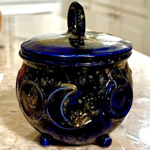 May include: A deep blue ceramic lidded jar with a glossy finish and gold-toned speckling. The jar features a rounded body, three small feet, and a lid with a central loop handle. Embossed moon phase symbols are visible on the exterior surface.