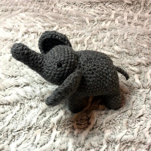 May include: A gray crocheted elephant toy with black eyes. The elephant is standing on a white fluffy surface.