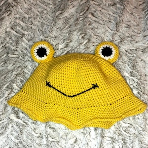 May include: A yellow crocheted frog hat with black eyes and a white outline. The hat has a wide brim and a smiling mouth.