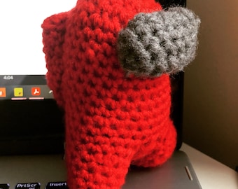 May include: A handmade, red crochet figure with a gray backpack-like detail. The figure is positioned on a laptop keyboard. The item is a representation of a character from the popular video game, Among Us.