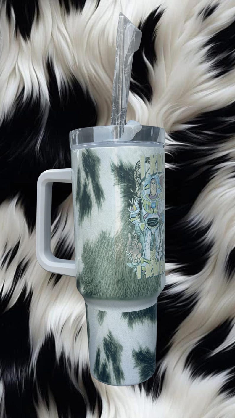 K&ouml;nnte beinhalten: Ein wei&szlig;er Becher mit gr&uuml;nem Kuhfleckenmuster, einem stabilen Griff, einem durchsichtigen Deckel und einem Kunststoffstrohhalm. Der Becher zeigt eine Grafik eines Weltraum-Ranger-Charakters, umgeben von floralen Elementen und dem Text I KINDA FRY.