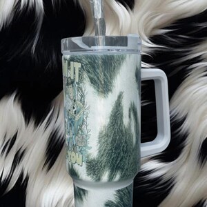 K&ouml;nnte beinhalten: Ein Edelstahlbecher mit einem wei&szlig;-dunkelgr&uuml;nen Kuhfellmuster, ausgestattet mit einem transparenten Deckel, einem wei&szlig;en Henkel und einem durchsichtigen Strohhalm. Der Becher zeigt eine grafische Illustration einer Cowboy-Figur.