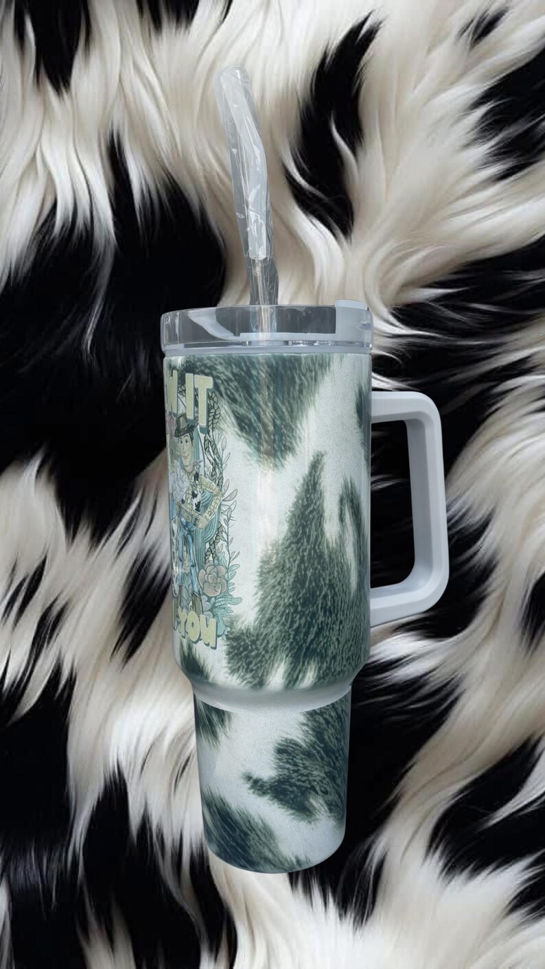 K&ouml;nnte beinhalten: Ein Edelstahlbecher mit einem wei&szlig;-dunkelgr&uuml;nen Kuhfellmuster, ausgestattet mit einem transparenten Deckel, einem wei&szlig;en Henkel und einem durchsichtigen Strohhalm. Der Becher zeigt eine grafische Illustration einer Cowboy-Figur.