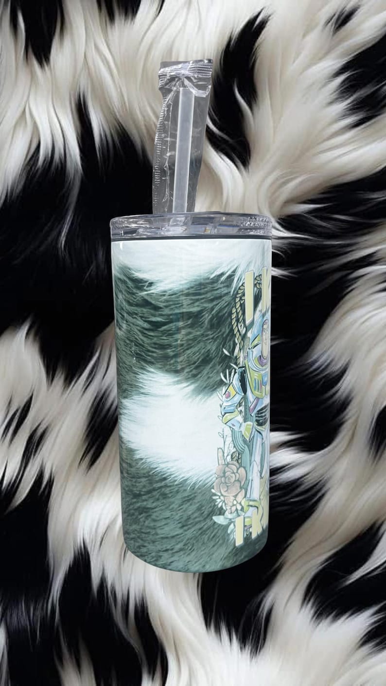 K&ouml;nnte beinhalten: Ein zylindrischer Edelstahlbecher mit transparentem Deckel und verpacktem Strohhalm. Die Au&szlig;enseite zeigt ein gr&uuml;n-wei&szlig;es Kuhfellmuster mit einer grafischen Illustration eines Weltraum-Rangers, floralen Akzenten und gelbem Text.