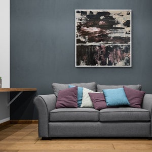 Może przedstawiać: Szara,  tkana sofa z czterema poduszkami w r&oacute;żnych odcieniach fioletu, niebieskiego i białego. Sofa stoi przed ciemnoszarą ścianą z abstrakcyjnym obrazem w ramie nad nią.