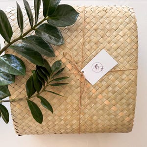 May include: A square woven basket with a tag tied with twine, and a sprig of green leaves. The basket is made of light brown woven material. The tag has a logo with the letter "B" and a leaf design.