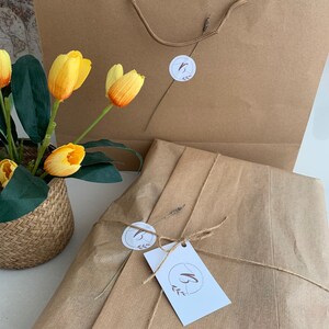 May include: Kraft paper shopping bag with a twisted handle and a wrapped package tied with twine. The package has a white tag with a logo. Yellow and orange tulips in a woven basket are in the foreground.