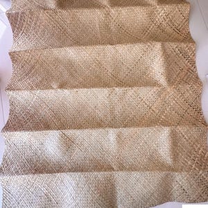 May include: A woven mat made of natural fibers, featuring a light brown color and a textured, rectangular design. The mat is folded in a zig-zag pattern, creating a layered effect. The mat is likely used for decorative or functional purposes.