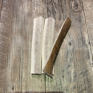 May include: Three natural antler dog chews, two are light brown and one is a darker brown. The chews are on a wooden surface.