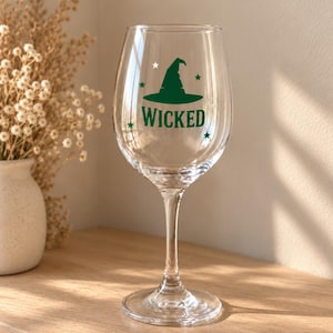May include: A clear glass wine glass featuring a green graphic of a witch hat and the word WICKED printed in green lettering. The design includes small green star accents surrounding the hat and text, positioned on a long, slender stem.
