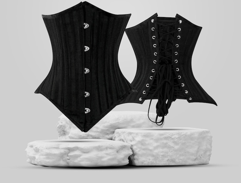 May include: A black corset with a busk closure and lace-up back. The corset is displayed on a white stone platform.