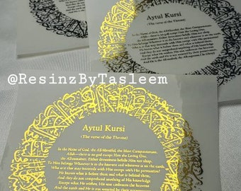 May include: A white card with gold calligraphy and the text "Aytul Kursi" (The verse of the Throne) in black text. The text is in English and Arabic. The card is on a gray surface.