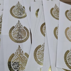 May include: A stack of white paper stickers with gold foil calligraphy. The calligraphy is in Arabic and reads "Allah".