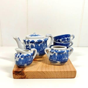 May include: A miniature tea set featuring a white teapot, a creamer, a sugar bowl, and two teacups, all decorated with a blue willow pattern. The set includes a lidded teapot with a curved handle and spout, a small creamer, and a two-handled sugar bowl.
