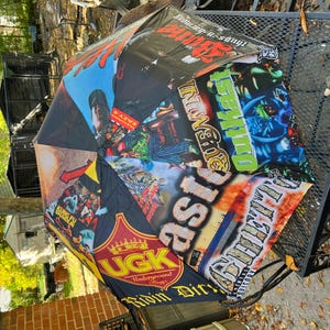 May include: A black umbrella with a vibrant collage of graphics and text. The design includes various logos, album art, and text elements in multiple colors. The umbrella is open, showcasing its full design.