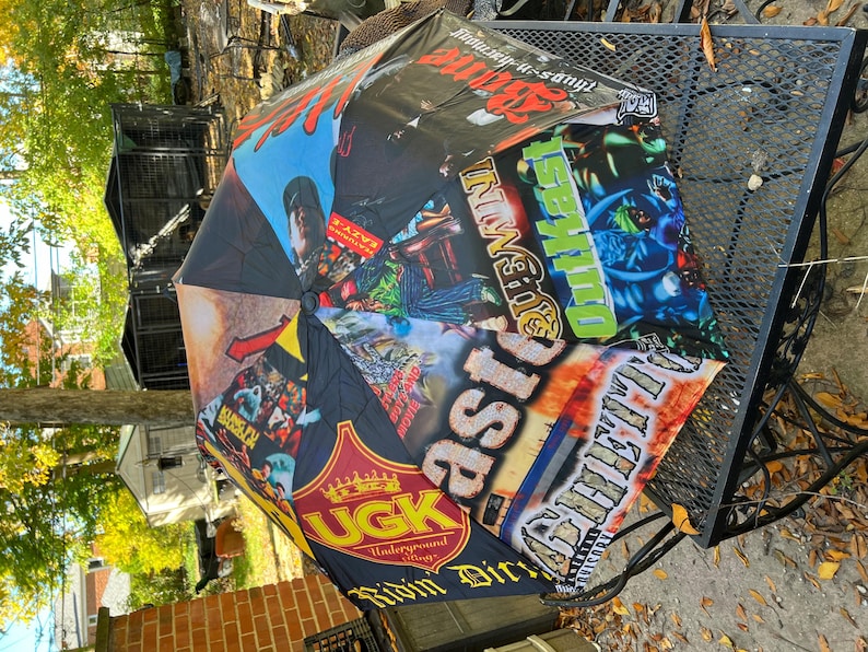 May include: A black umbrella with a vibrant collage of graphics and text. The design includes various logos, album art, and text elements in multiple colors. The umbrella is open, showcasing its full design.