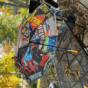 May include: Black umbrella with a variety of colorful graphics and text. The umbrella is open, displaying a collage of images and text. The handle is brown with a curved design. The umbrella is resting on a metal table.