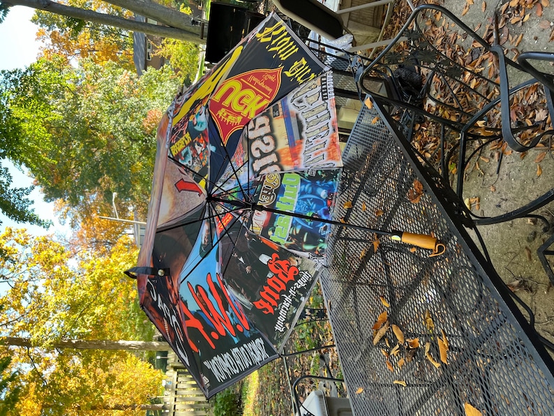 May include: Black umbrella with a variety of colorful graphics and text. The umbrella is open, displaying a collage of images and text. The handle is brown with a curved design. The umbrella is resting on a metal table.