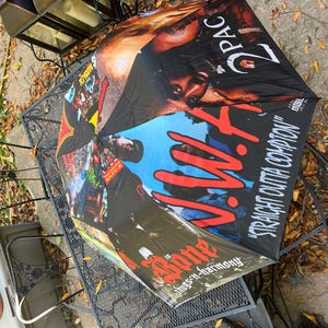 May include: Black umbrella featuring images of 2Pac and N.W.A. The umbrella has a black background with the words "Straight Outta Compton" and "Bone thugs-n-harmony" printed on it. The umbrella is open and resting on a black metal table.