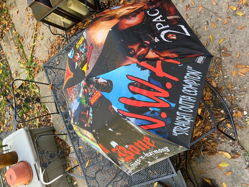 May include: Black umbrella featuring images of 2Pac and N.W.A. The umbrella has a black background with the words "Straight Outta Compton" and "Bone thugs-n-harmony" printed on it. The umbrella is open and resting on a black metal table.