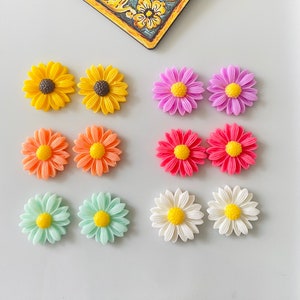 Può includere: Assortimento di fiori colorati in resina. I fiori sono di vari colori, tra cui giallo, arancione, rosa, viola, bianco e verde. I fiori sono disposti a coppie su una superficie bianca.