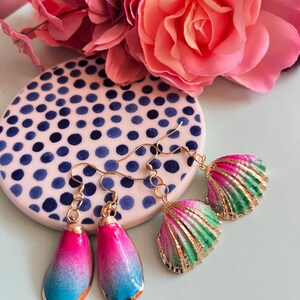 May include: Two pairs of earrings with gold-colored hooks. One pair features teardrop-shaped pendants with a gradient of pink, blue, and orange. The other pair has shell-shaped pendants with pink, green, and gold accents. A pink floral arrangement and a polka-dot coaster are in the background.