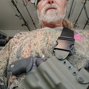 Könnte beinhalten: Ein mattes, olivgrünes, geformtes Holster mit schwarzen Beschlägen und einem schwarzen verstellbaren Riemen, das einen schwarzen Revolver mit strukturiertem Griff enthält. Das Holster wird über einem langärmeligen Shirt mit Tarnmuster getragen.
