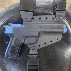 May include: Black polymer holster with a Ruger pistol. The holster features three black buckles and a strap. The pistol is black with blue tape on the front. The holster is designed to be worn on the leg.