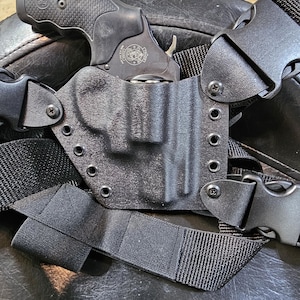 Könnte beinhalten: Ein schwarzes Lederholster mit einer schwarzen Pistole darin. Das Holster ist an einem schwarzen Schultergurt mit verstellbaren Schnallen befestigt.