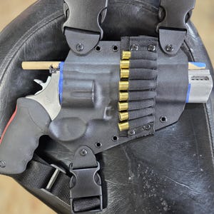 May include: A black moulded holster featuring a side-mounted elastic strip holding eight brass-coloured cartridges. The holster includes adjustable black nylon straps with plastic side-release buckles and contains a silver-toned revolver.