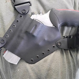 May include: A black molded holster with black nylon straps and plastic side-release buckles. The holster contains a silver revolver with a black and red grip. The holster features a series of metal eyelets along the edge and is secured with screws.