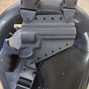 May include: A black holster with a black strap and buckle, holding a silver and black revolver with a blue grip. The holster has the number "686" written on it. The holster is designed to be worn on the leg.