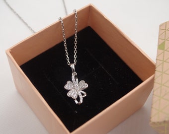 Puede incluir: Un collar de plata con un colgante de trébol de cuatro hojas engastado con piedras de circonita cúbica transparentes. El collar está en una caja de regalo rosa claro con un forro de terciopelo negro.