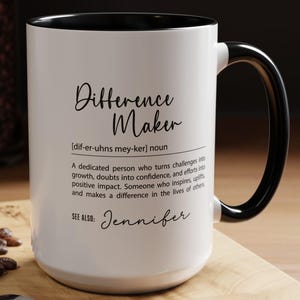 May include: White ceramic coffee mug with a black rim and handle. The mug has black text that reads "Difference Maker" with the phonetic pronunciation and definition below. The text "See Also: Jennifer" is printed below the definition.