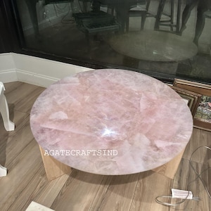 May include: A round coffee table with a light pink, rose quartz top. The table has a natural wood base and is set on a light wood floor. The text "AGATECRAFTSIND" is visible on the table.