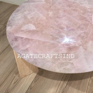 May include: A round coffee table with a rose quartz top. The table has a light pink, textured surface and is supported by a light-colored wooden base. The text "AGATECRAFTSIND" is visible on the table.