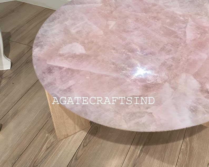 May include: A round coffee table with a rose quartz top. The table has a light pink, textured surface and is supported by a light-colored wooden base. The text "AGATECRAFTSIND" is visible on the table.
